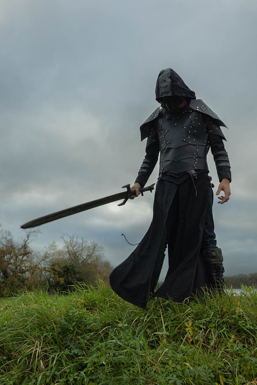 A fantasy figure in a long black cloak  stands in a field holding a sword.